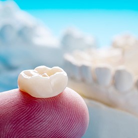 Dental crown resting on a fingertip in front of plaster jaw with light blue background